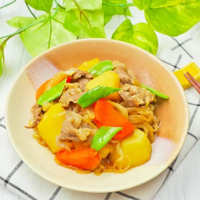 お皿に盛られた肉じゃが。ジャガイモ、ニンジン、グリーンピース、牛肉が使われています。後ろに緑の葉が見えます。