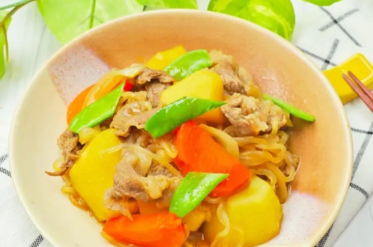 肉じゃがが皿に盛られている。にんじん、じゃがいも、さやえんどうなどの野菜と肉が見える。背景には観葉植物。