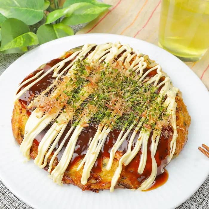 長ねぎとツナのお好み焼きが皿にのっている。上にマヨネーズ、ソース、青のり、かつお節がかかっている。背景には飲み物がある。