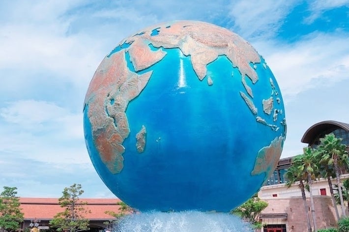 大きな地球儀のオブジェが噴水の中に置かれ、背景には青空と建物があります。