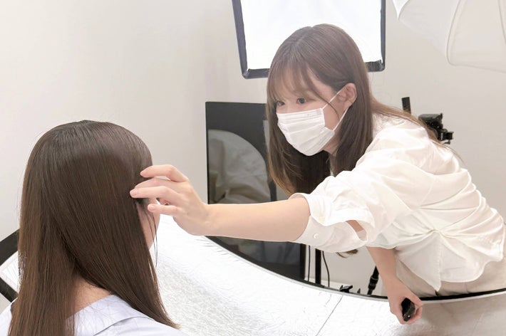 女性が白い背景の前に立っている。力を抜いた優しい表情を見せ、スタイリッシュなシャツを着ている。