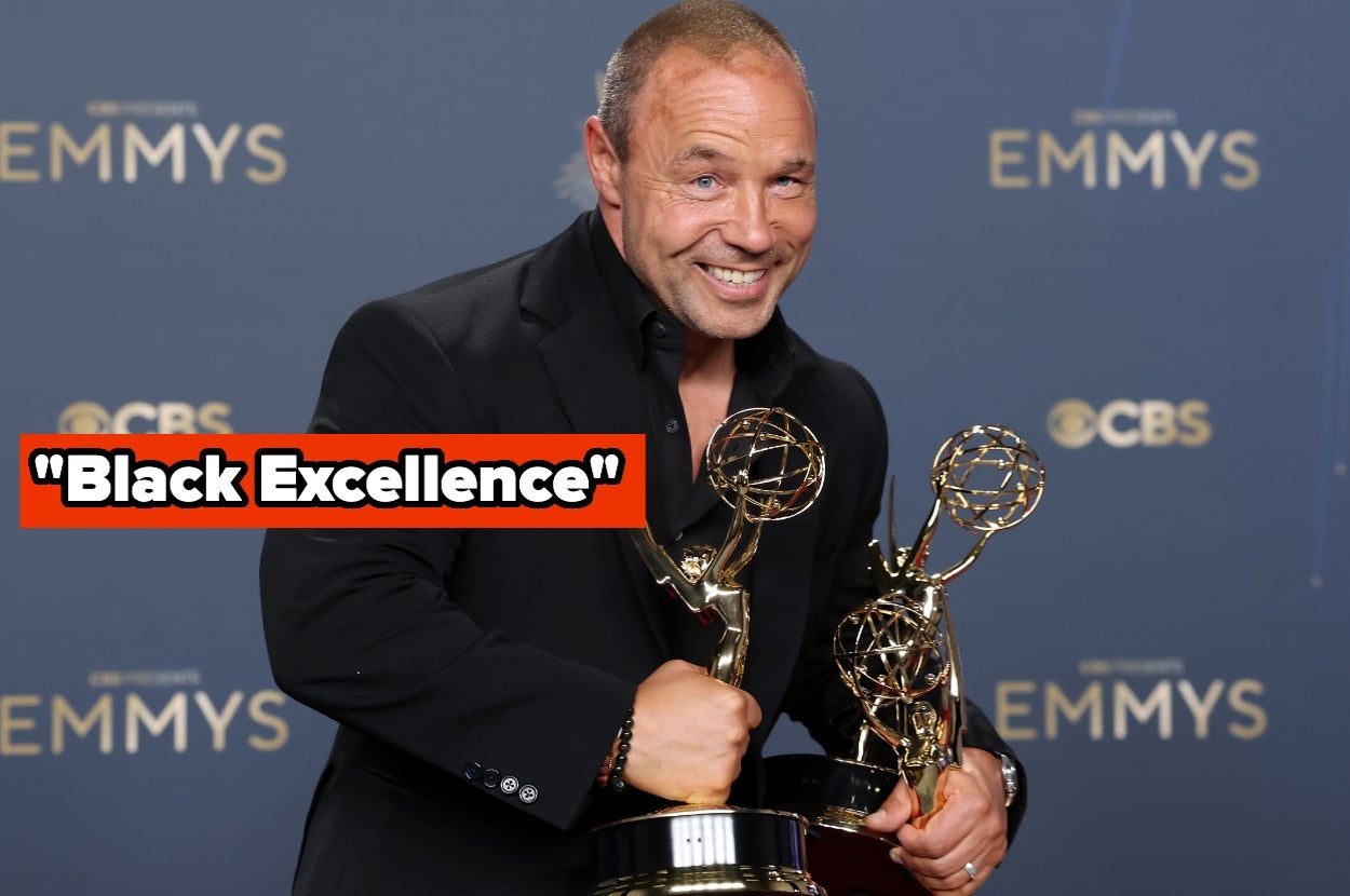 Man in a suit holding two Emmy awards, smiling at an event with "Emmys" signage in the background, text overlay reads "Black Excellence."