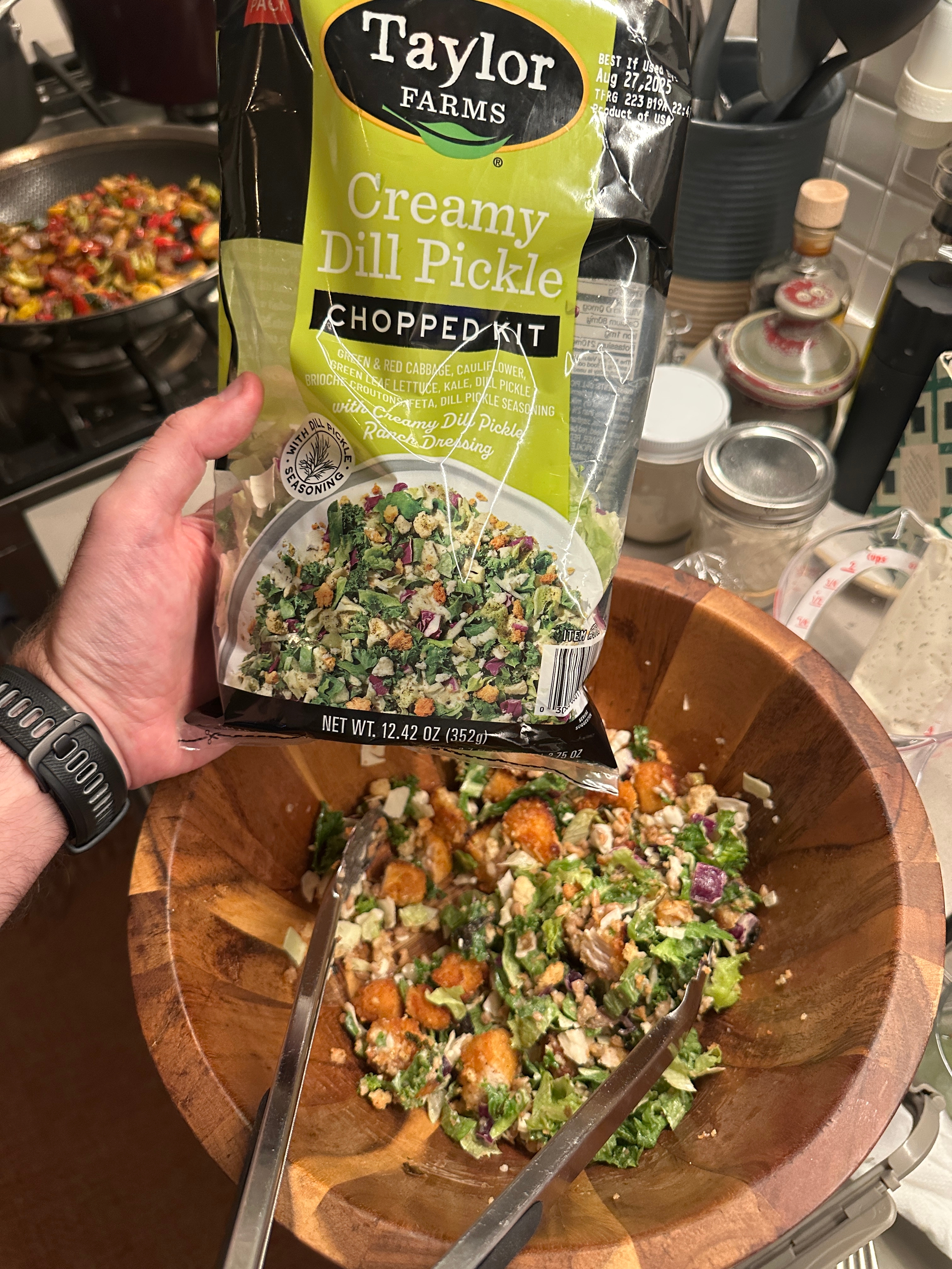 Bag of Taylor Farms Creamy Dill Pickle Chopped Kit held above a salad in a wooden bowl with tongs, in a kitchen setting