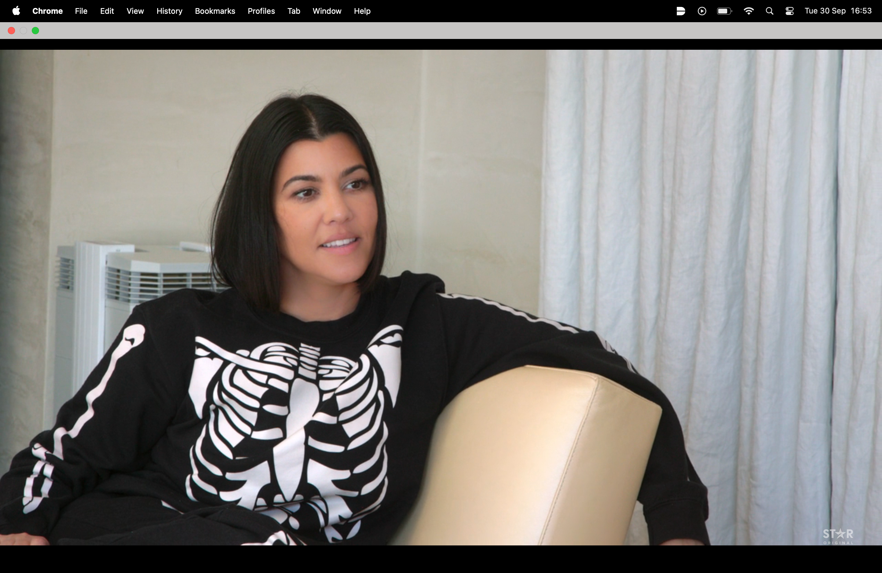 A person is sitting on a beige chair, wearing a casual outfit with a skeleton design on the top