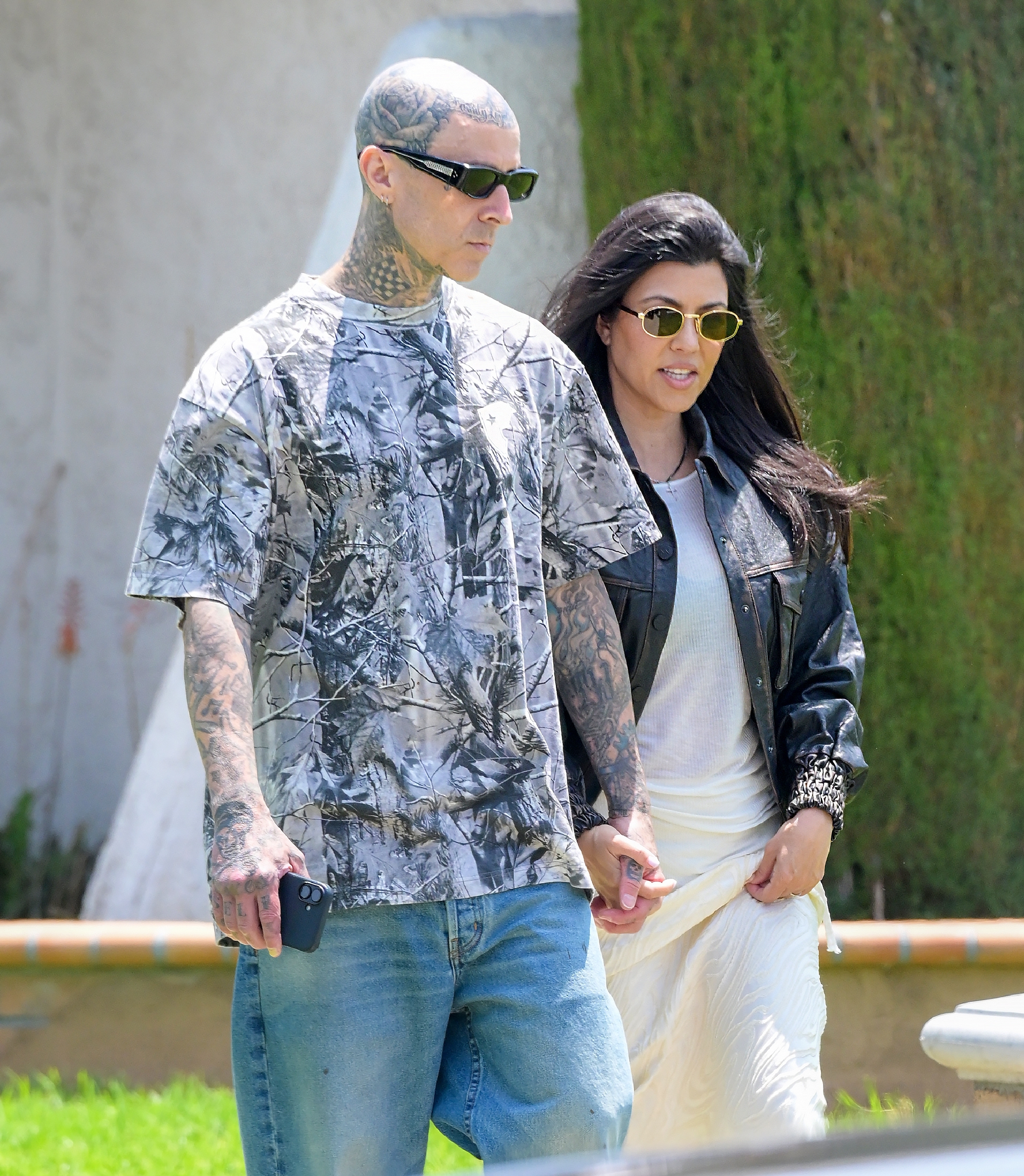 Two people walking hand-in-hand outdoors. The man wears a graphic t-shirt; the woman wears sunglasses, a leather jacket, and a light skirt