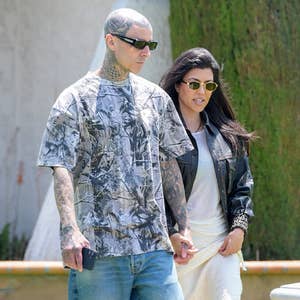 Two people walking hand-in-hand outdoors. The man wears a graphic t-shirt; the woman wears sunglasses, a leather jacket, and a light skirt