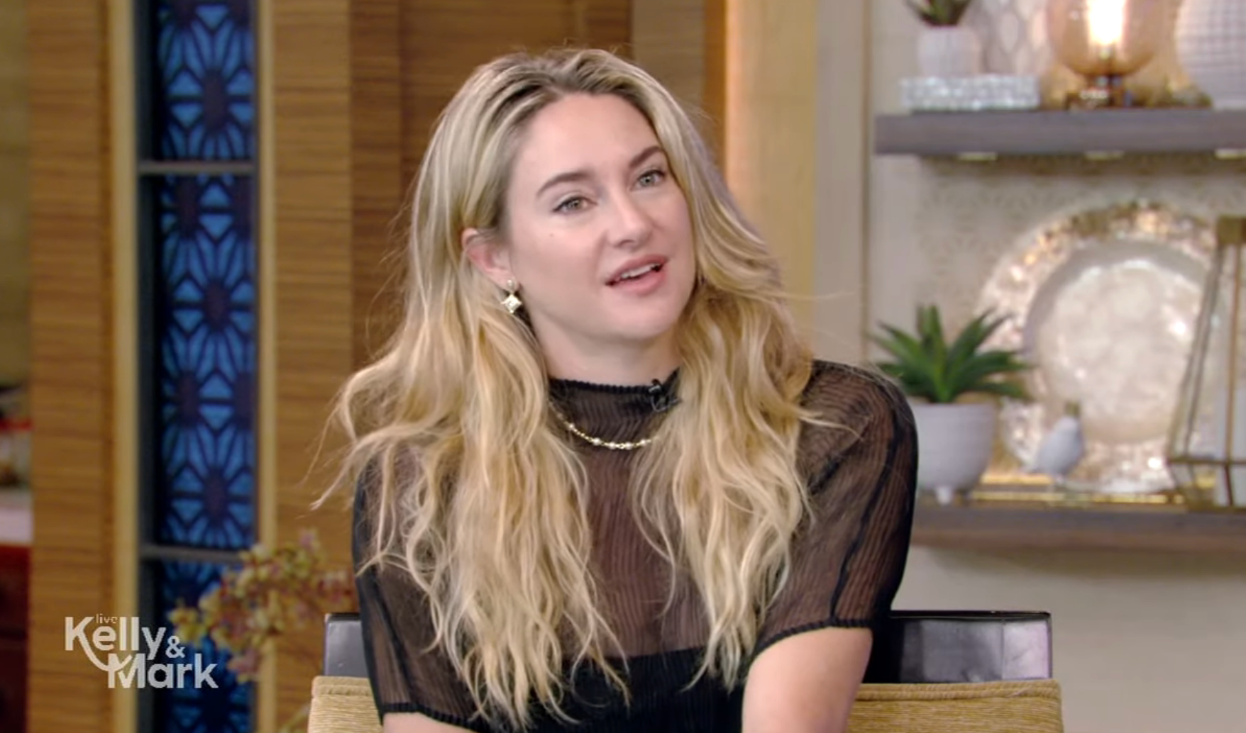 Person with long hair in a sheer, black top with delicate patterns, sitting on a talk show set. The atmosphere is casual and conversational