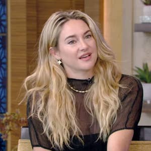 Person with long hair in a sheer, black top with delicate patterns, sitting on a talk show set. The atmosphere is casual and conversational