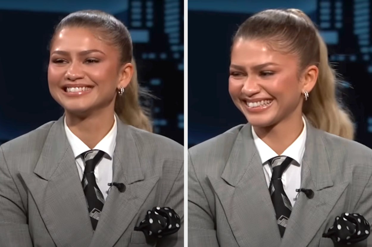 Person in a tailored suit with a tie and polka dot pocket square, smiling during a talk show appearance. Hair is styled in a ponytail