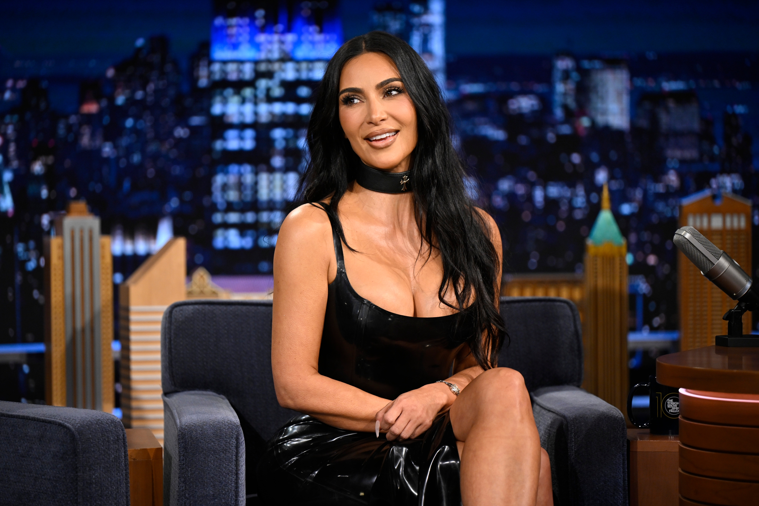 Person in a black leather dress sits on a talk show set, smiling during an interview