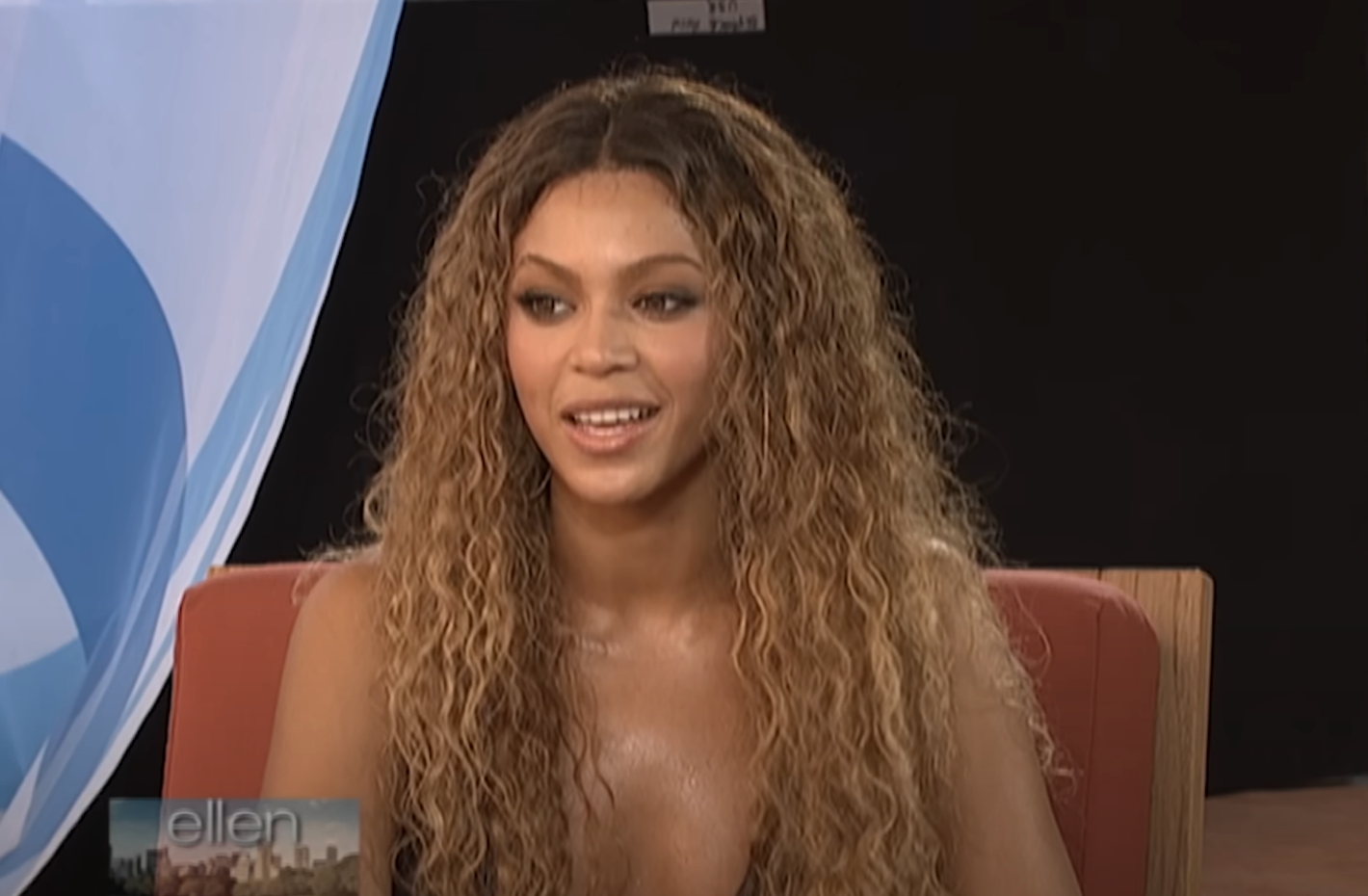 Celebrity with long wavy hair, wearing a low-cut top, smiling during an interview on a talk show set