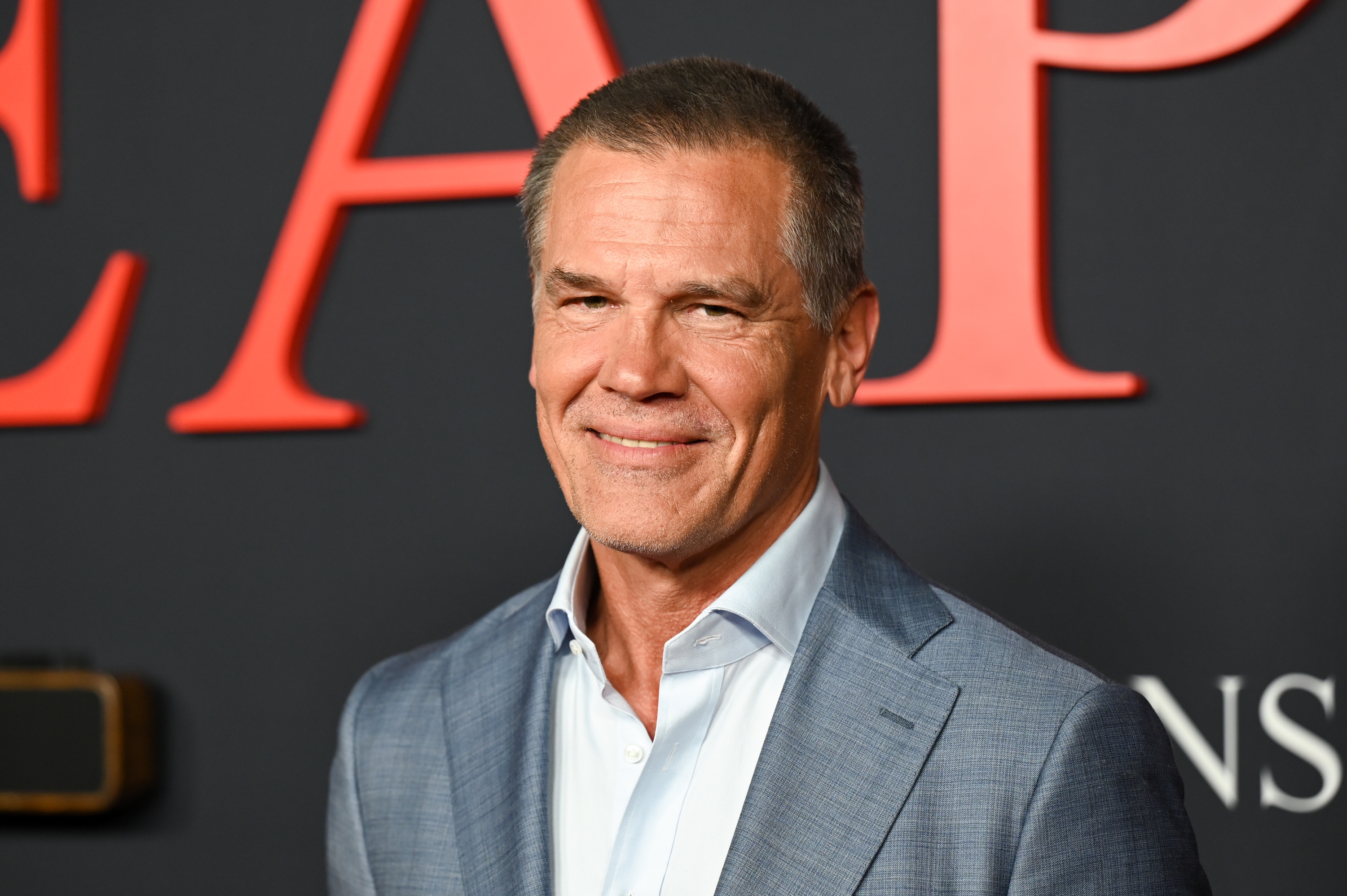 A man in a sharp suit smiles at a red carpet event with a backdrop featuring large letters