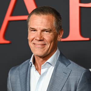 A man in a sharp suit smiles at a red carpet event with a backdrop featuring large letters