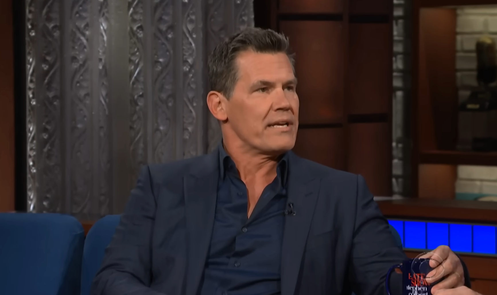 A man in a dark suit and shirt speaks while seated on a talk show set, holding a coffee mug