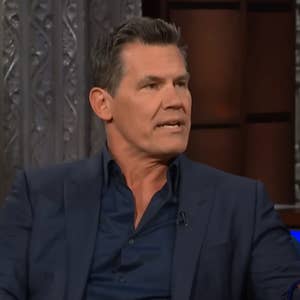 A man in a dark suit and shirt speaks while seated on a talk show set, holding a coffee mug