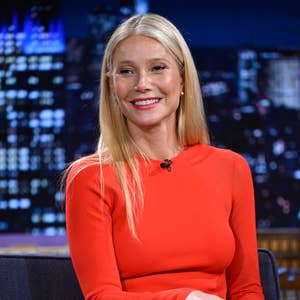 Person in long-sleeve gown sits on a talk show set, smiling during an interview, with a nighttime cityscape background