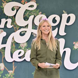 A celebrity smiles while holding a paper, standing in front of floral-themed "in Goop Health" sign, dressed in a casual, elegant outfit
