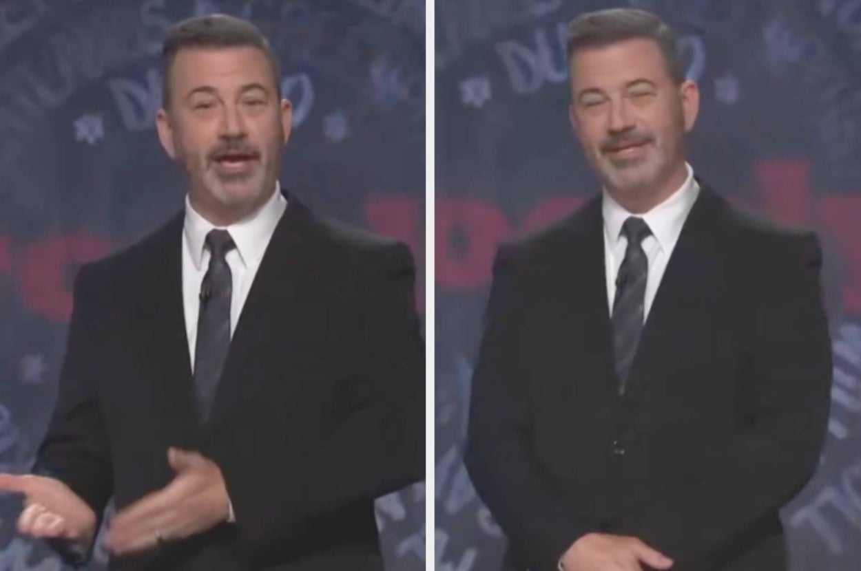 Person in a black suit and tie, gesturing and smiling, on stage with blurred text backdrop