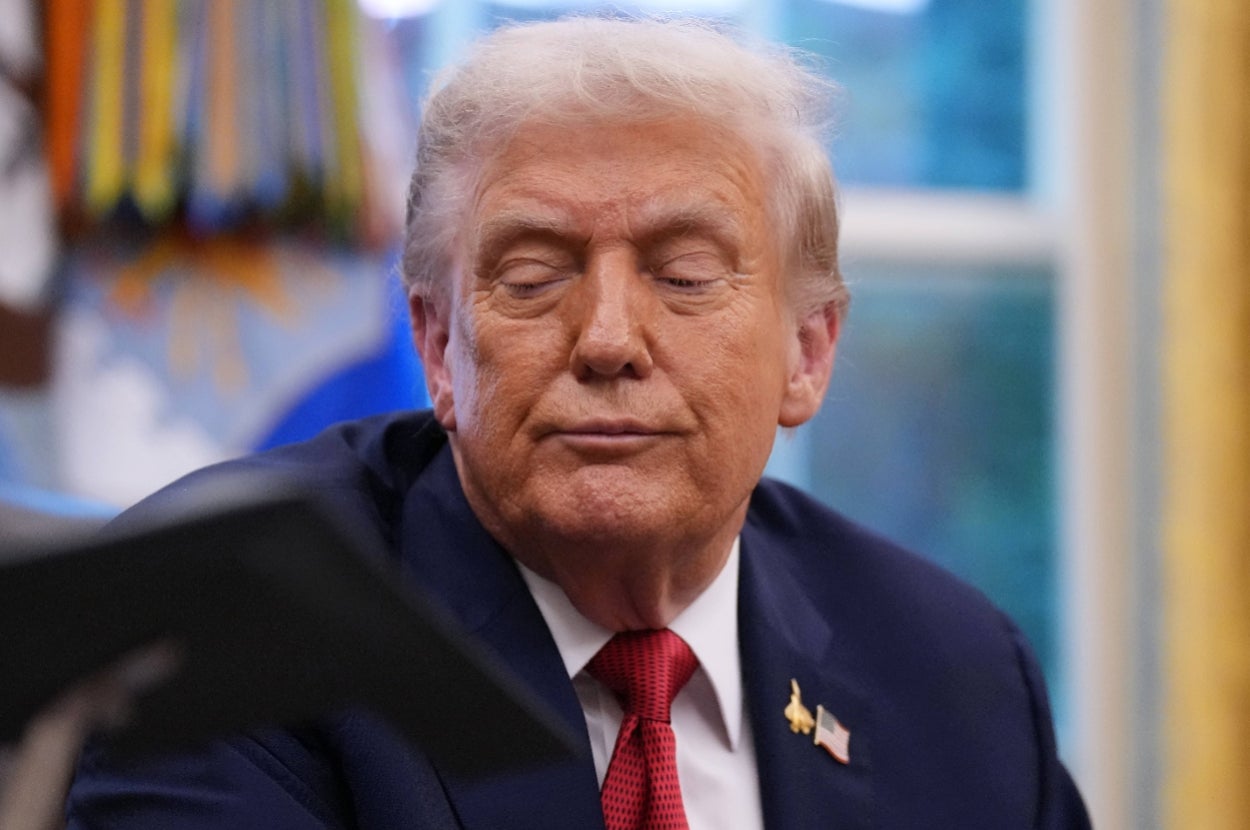 A person in a suit is sitting indoors, looking down with a thoughtful expression. Ribbons are visible in the background