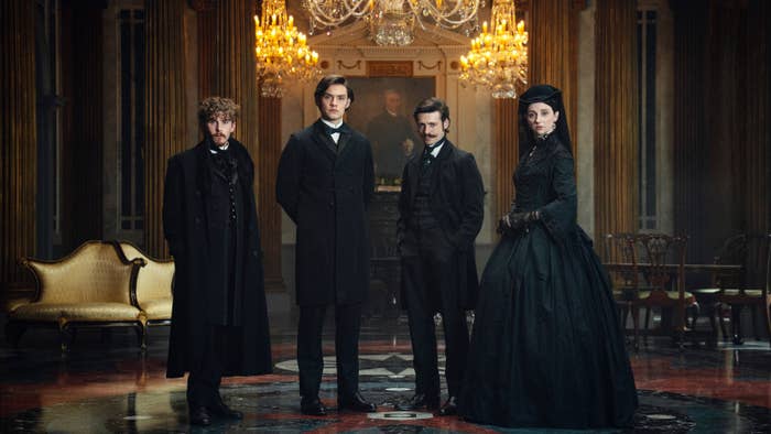 Period drama scene with four actors in 19th-century formal attire, set in an ornate room with chandeliers