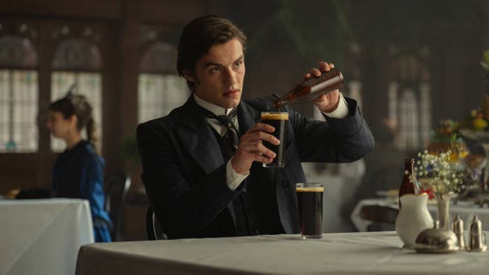 A man in period clothing pours a drink in a formal dining setting, suggesting a historical or dramatic film or TV scene