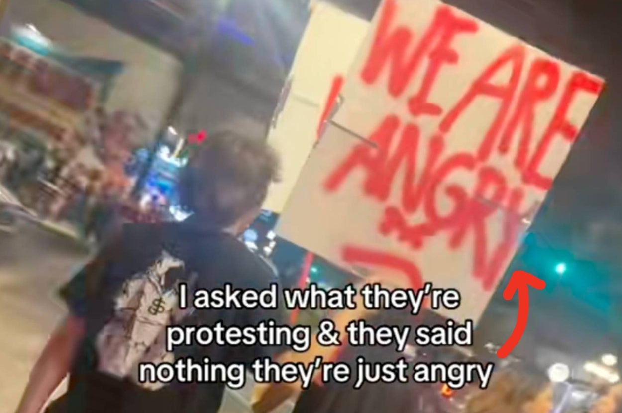 People holding signs reading "WE ARE ANGRY" and "NOT A PROTEST SIGN" on a street. The text overlay says they're not protesting, just angry