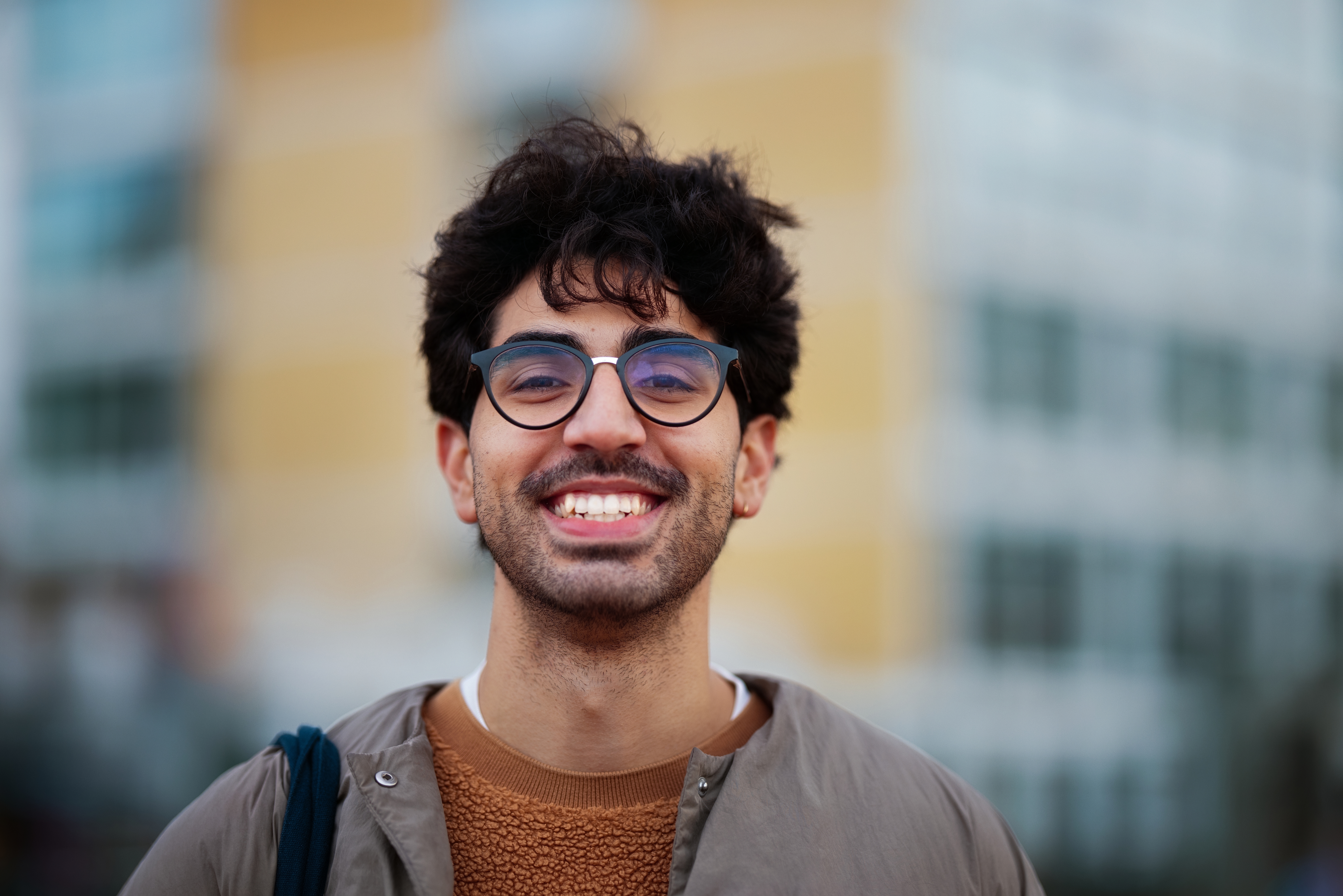 Person som smiler utendørs, iført briller og en uformell jakke over en genser. Urbane omgivelser er mykt uskarpe i bakgrunnen