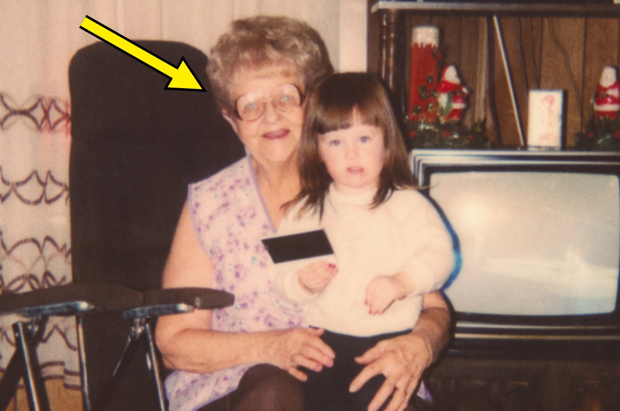 An older woman with glasses and a young child holding a photo sit in a living room; a yellow arrow points to the woman