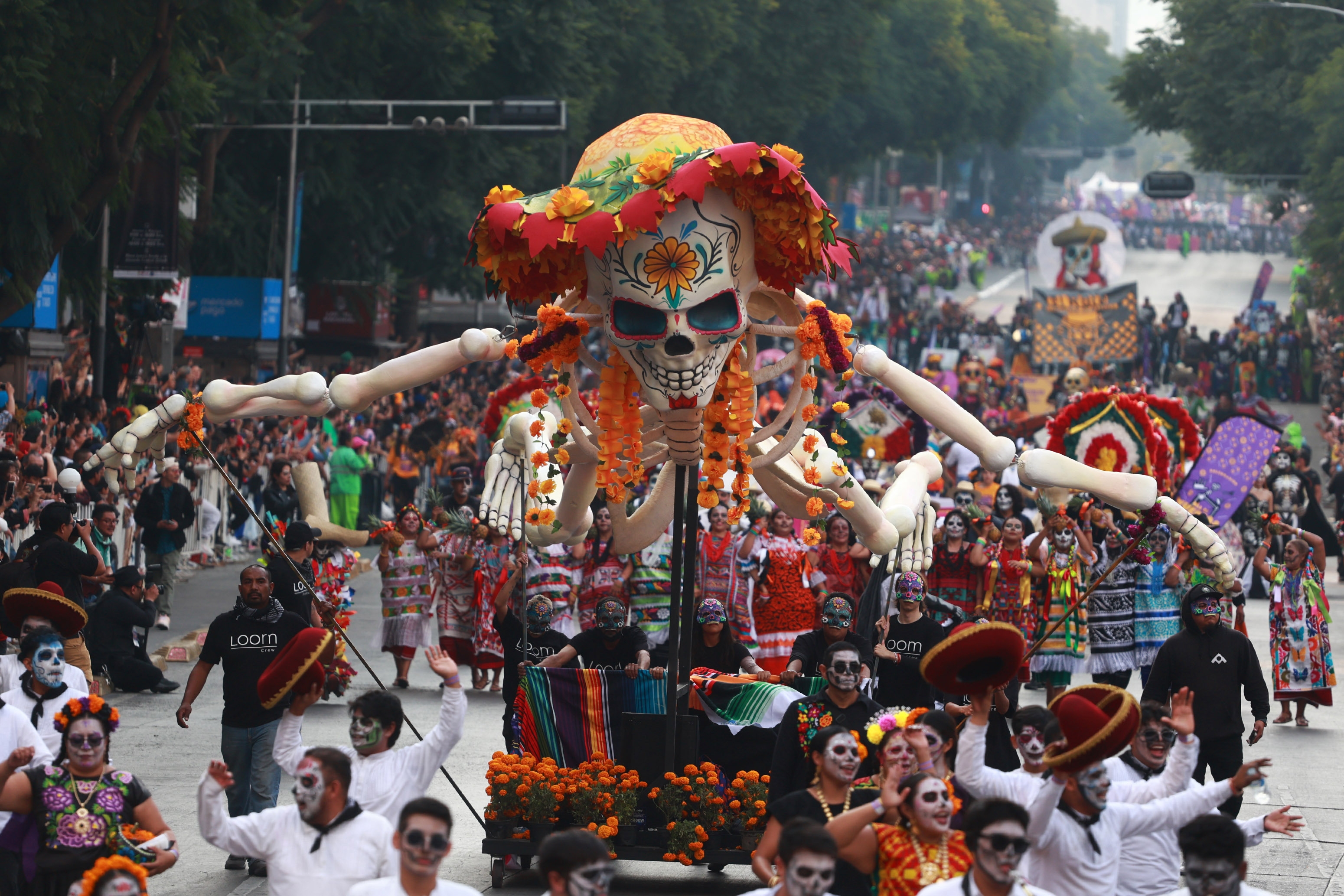 Parade with a giant skeleton float decorated with flowers and crowds dressed in traditional attire celebrating Dia de los Muertos on a lively street