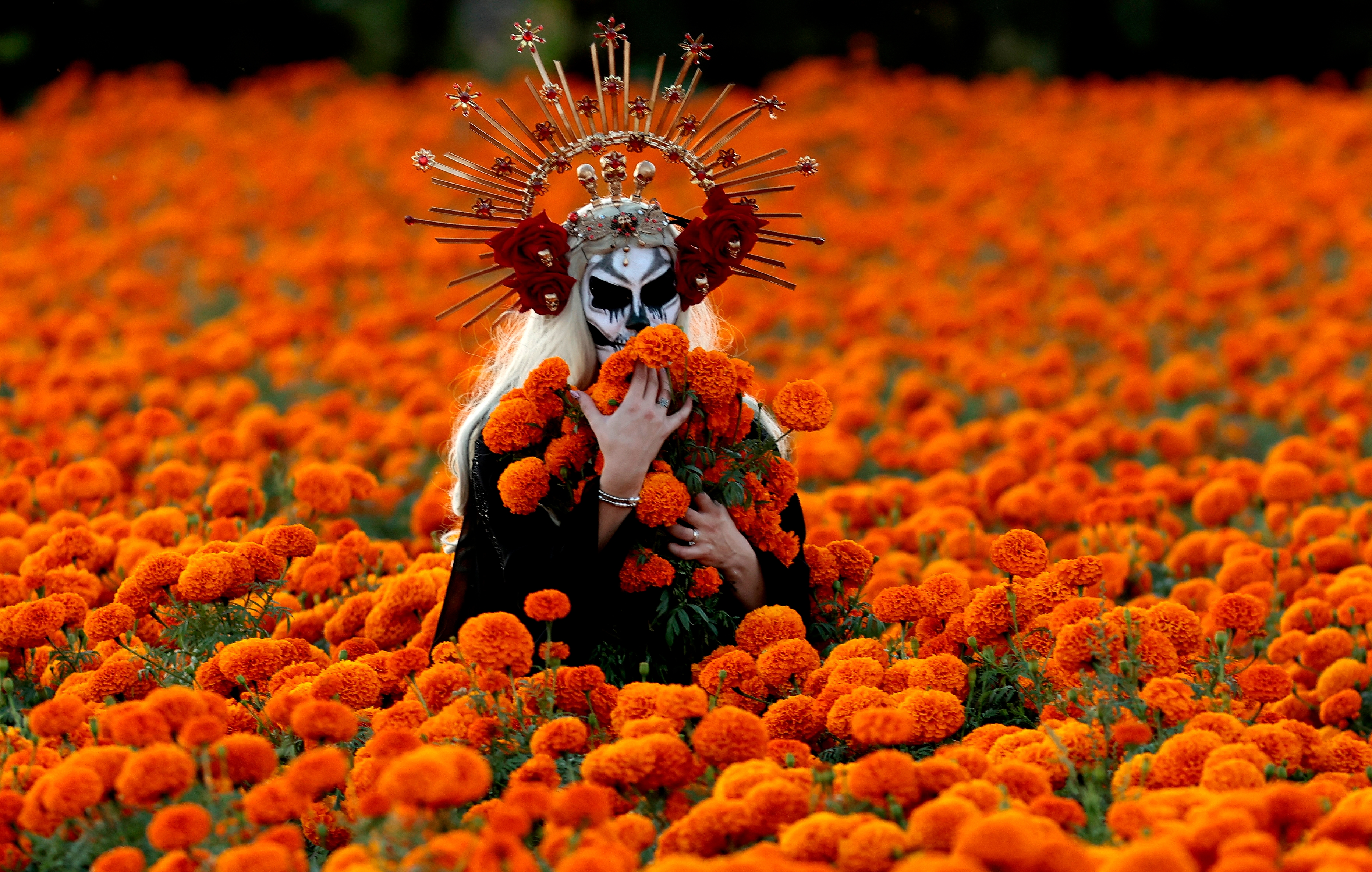 Person in ornate headdress and face paint stands in a marigold field, holding flowers