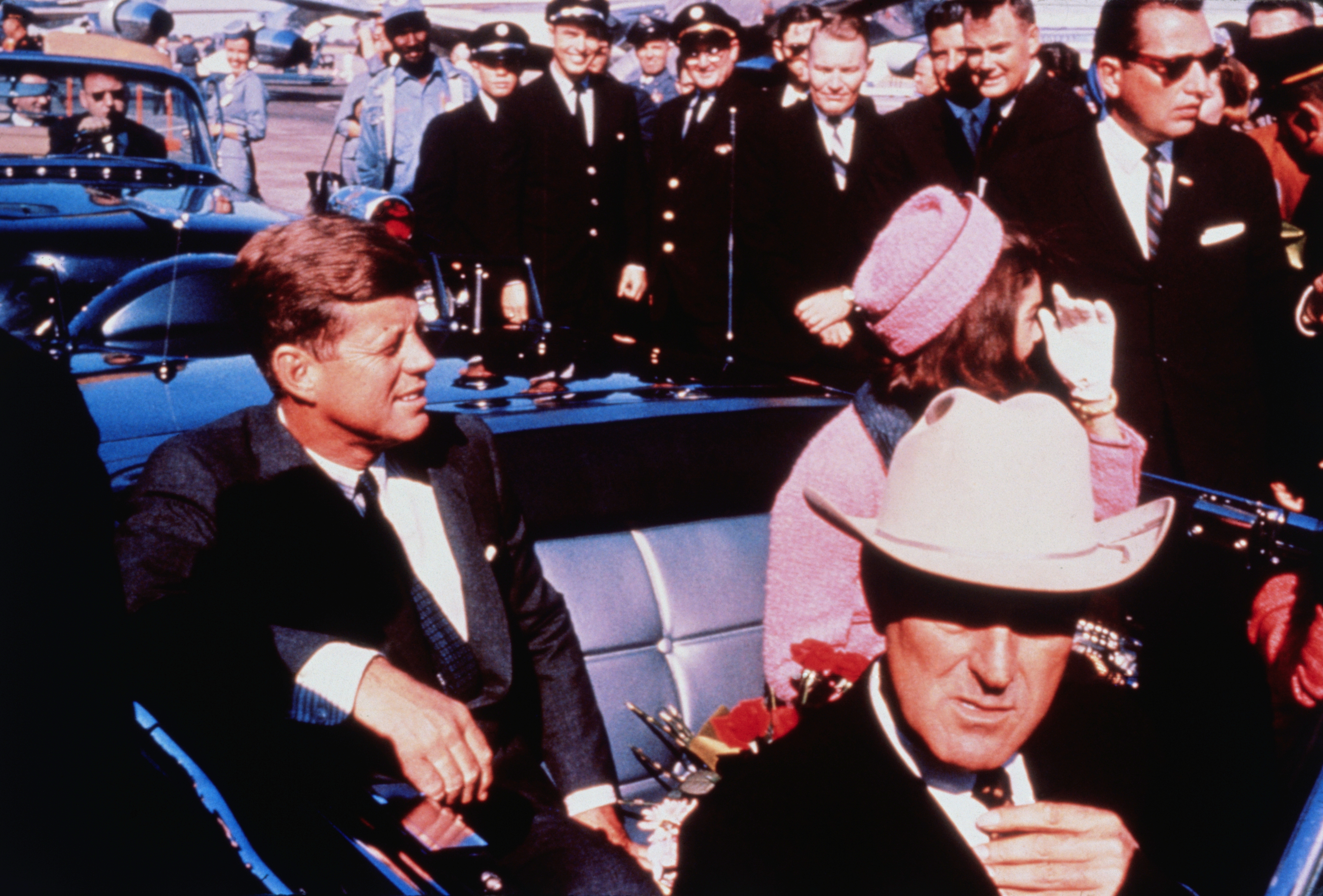 Historical photograph of President John F. Kennedy successful a motorcade with respective people, including a antheral successful a suit and a pistillate successful a hat. Crowds and officials are successful the background