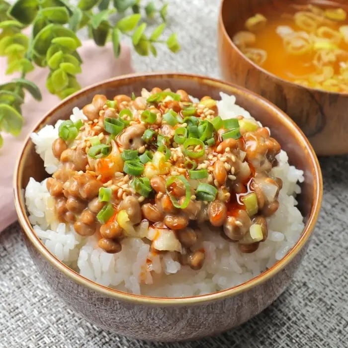 納豆ご飯が盛られた木の器。トッピングにはネギとごまが見える。バックには味噌汁が置かれ、食卓の一部を成している。