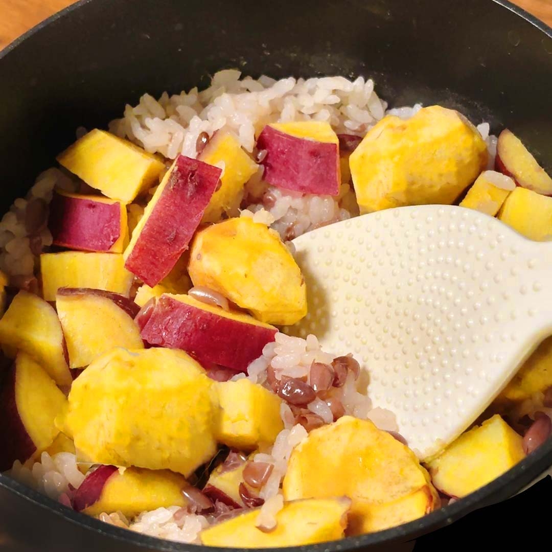炊かれたご飯とサツマイモが混ざり、白いしゃもじで混ぜている。