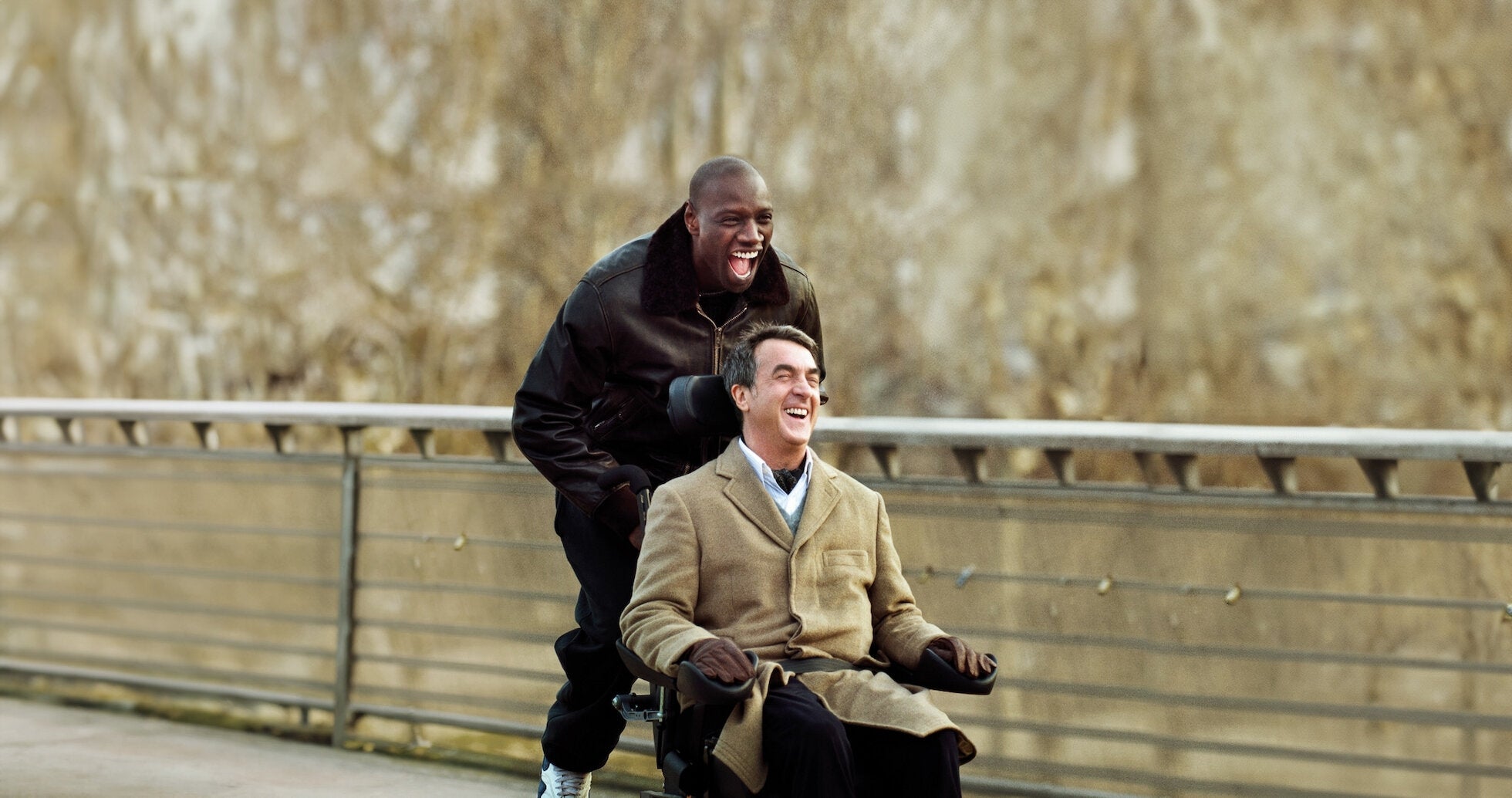 A man in a coat sits in a wheelchair, smiling, while another man in a jacket pushes him with joy. They are outdoors on a bridge