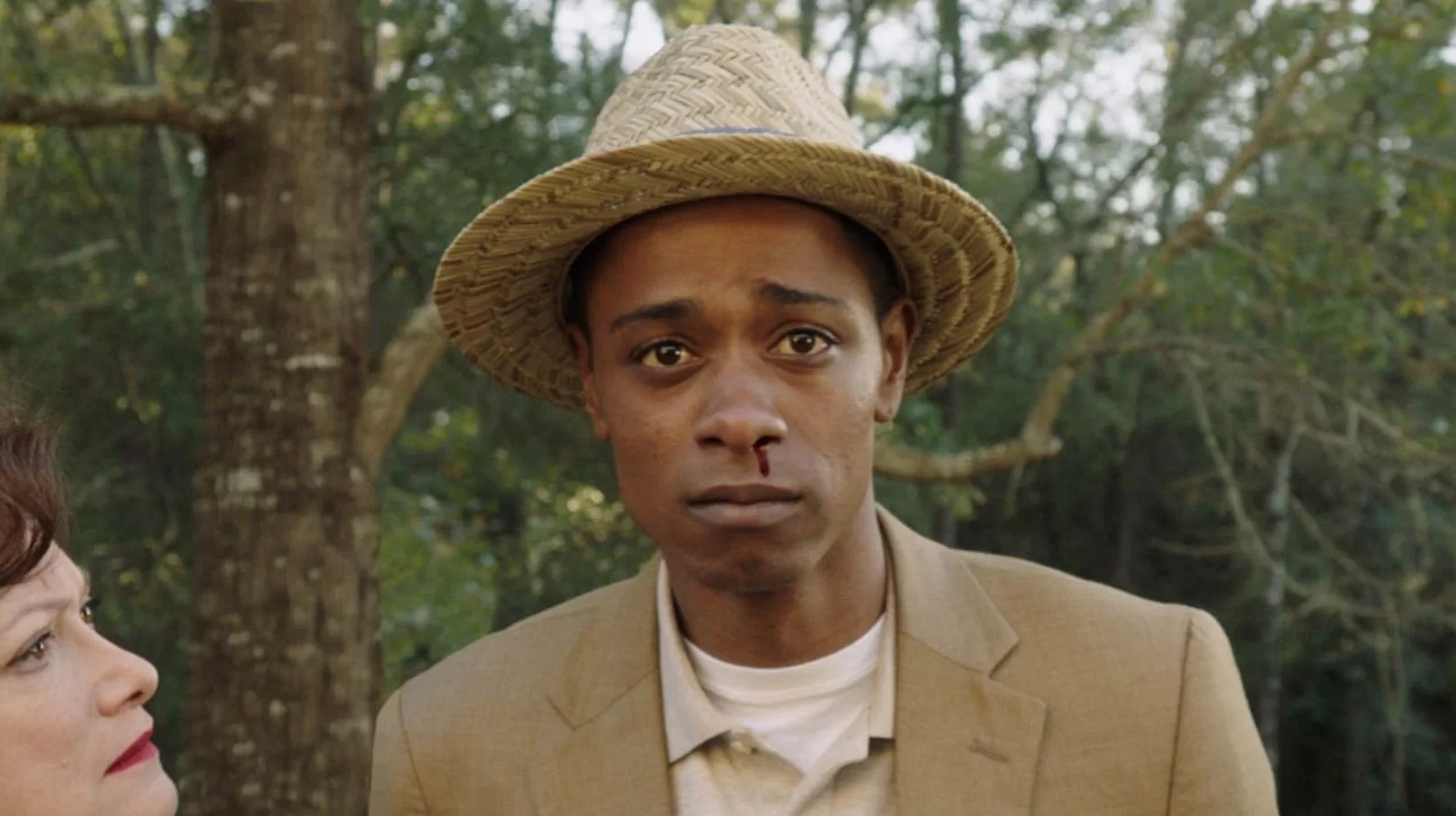 A person in a beige suit and straw hat stands outdoors, with a serious expression and a nosebleed, likely part of a TV or movie scene