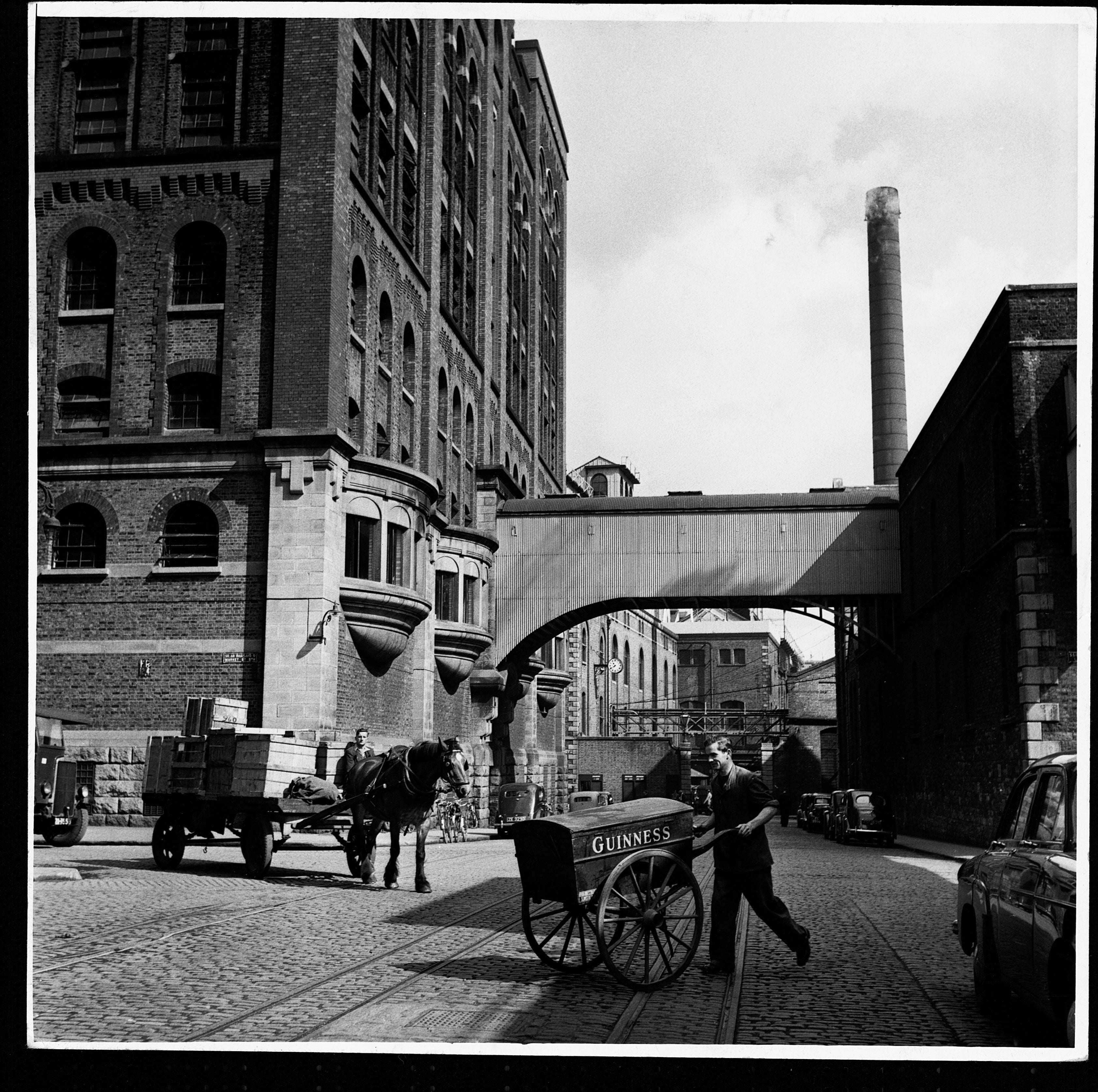 Historic image of a horse-drawn cart and a pedestrian near a brewery in an industrial cityscape