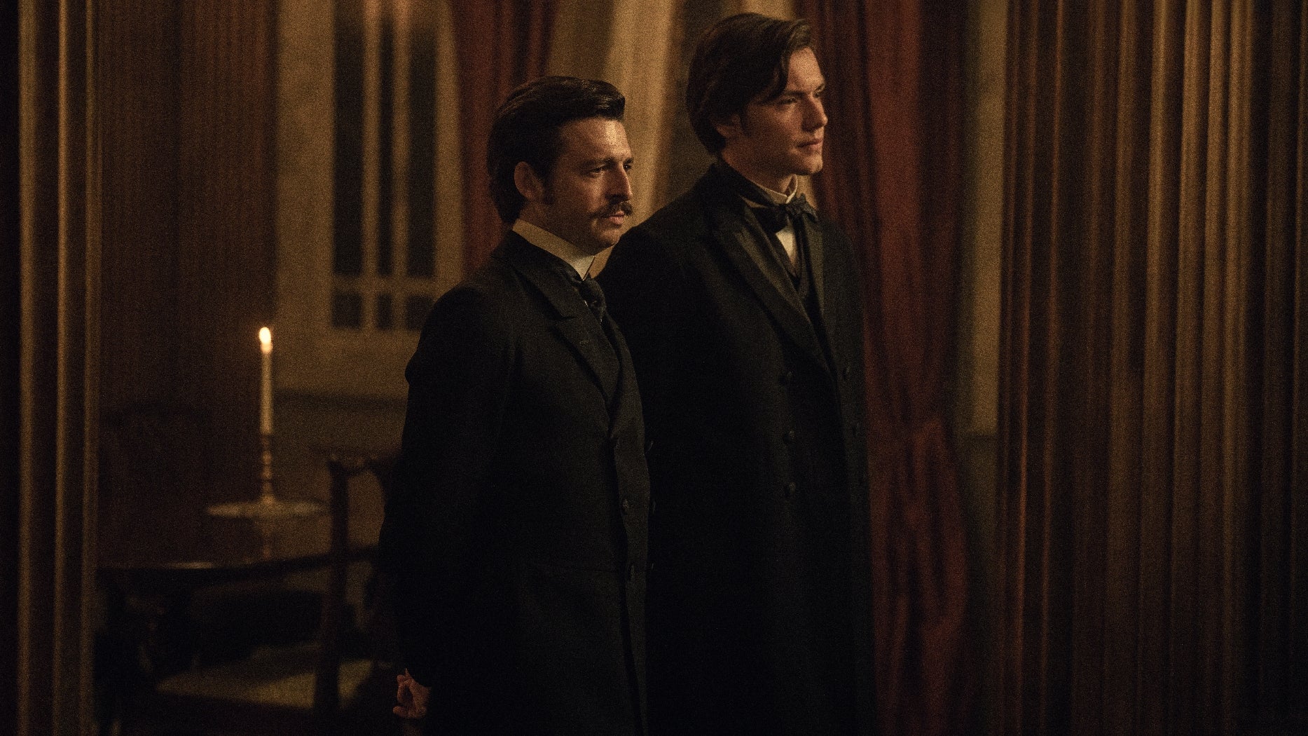 Two people in formal 19th-century suits stand in a dimly lit room with tall curtains and a lit candle in the background, evoking a historical setting