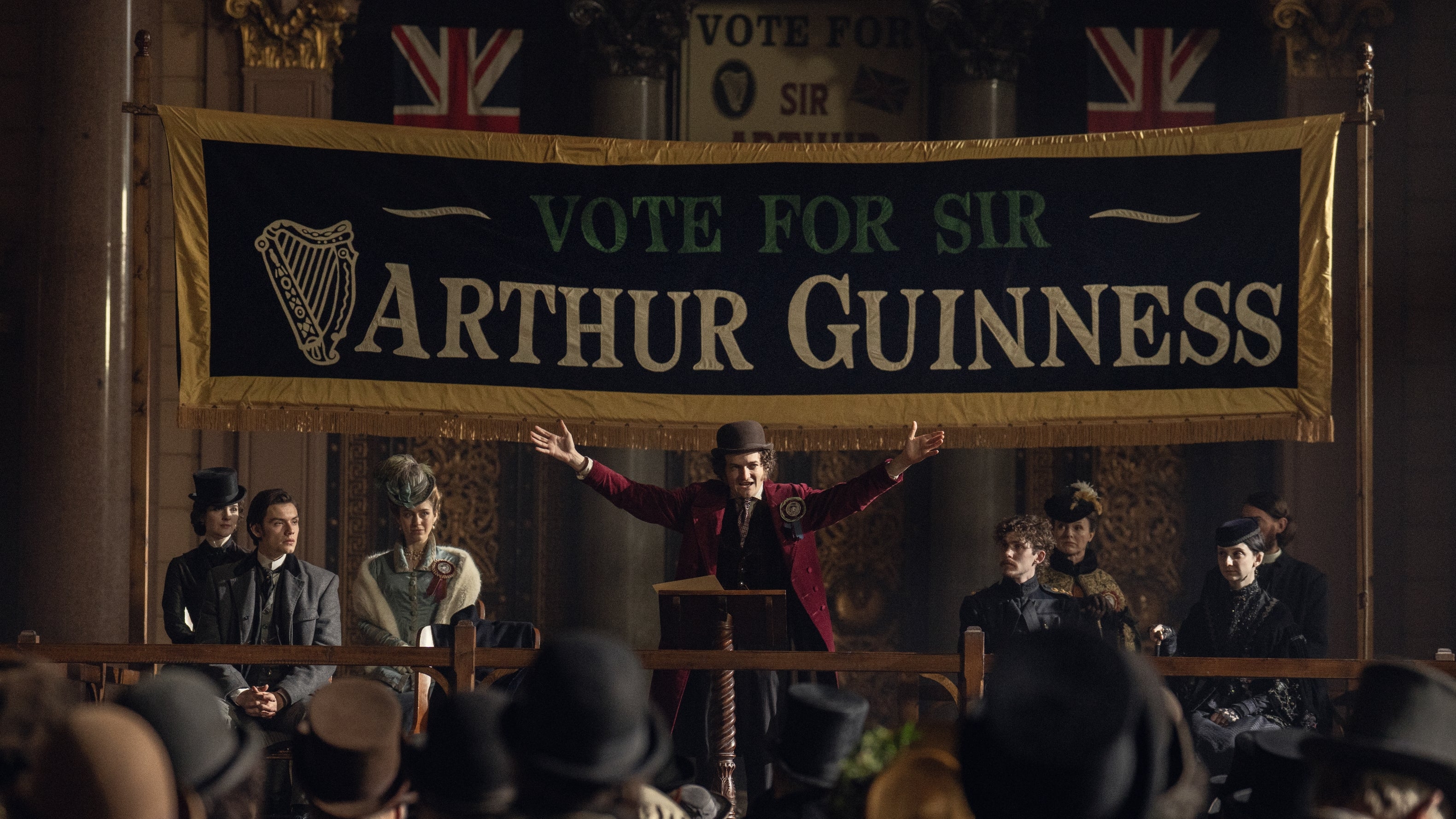 A character in historical attire, campaigning under a banner reading "Vote for Sir Arthur Guinness," addresses a formal assembly
