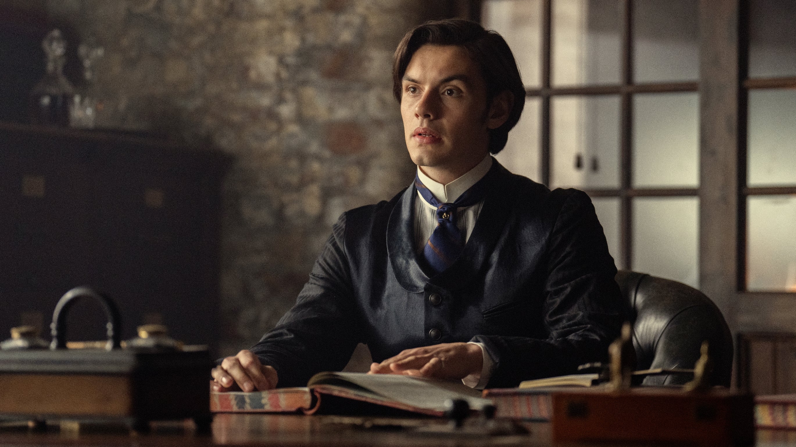 A person in period clothing sits at a desk in a dimly lit room, looking contemplative