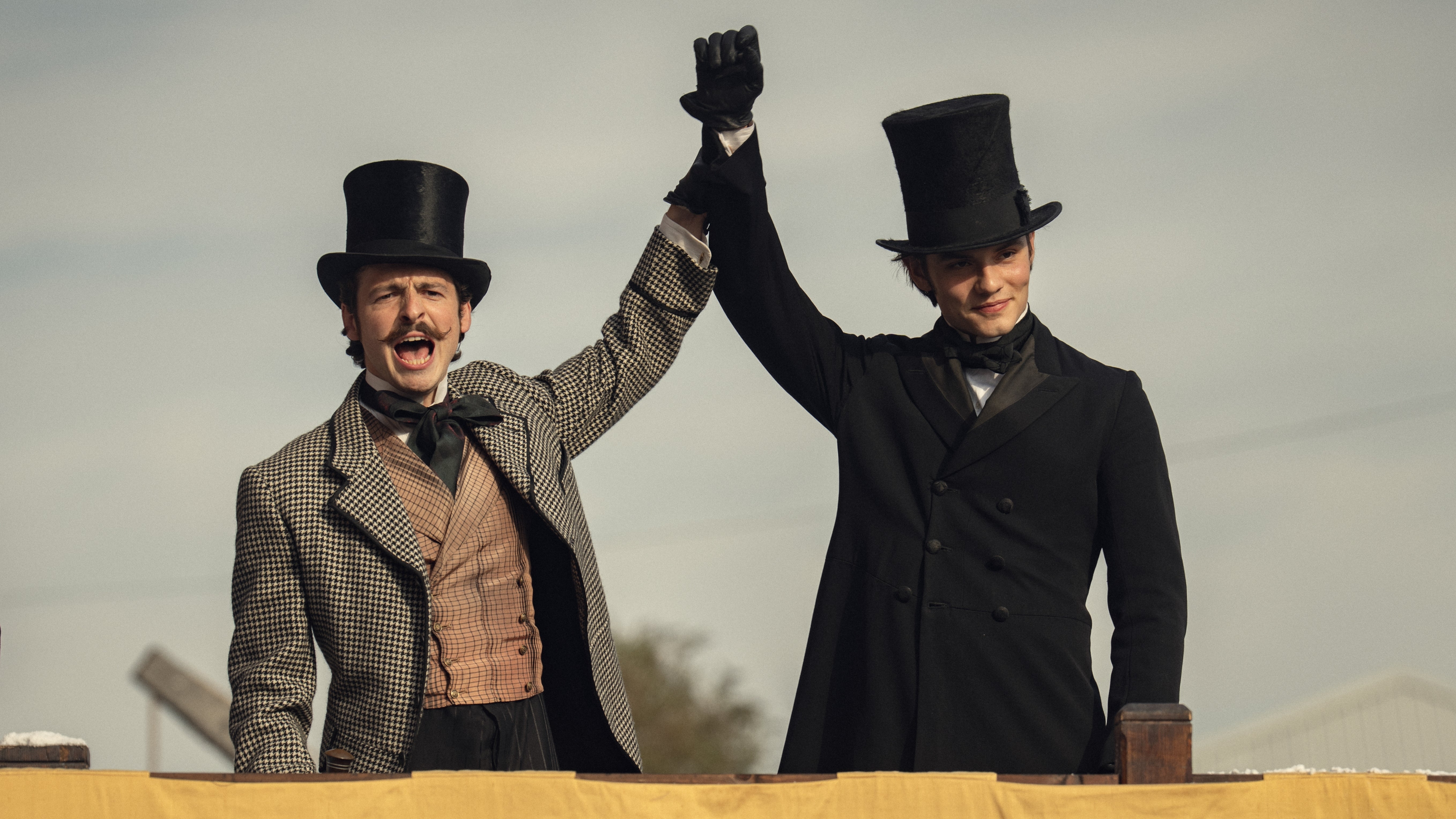 Two people in vintage attire and top hats stand on a platform, raising their clasped hands victoriously, reminiscent of a historical scene