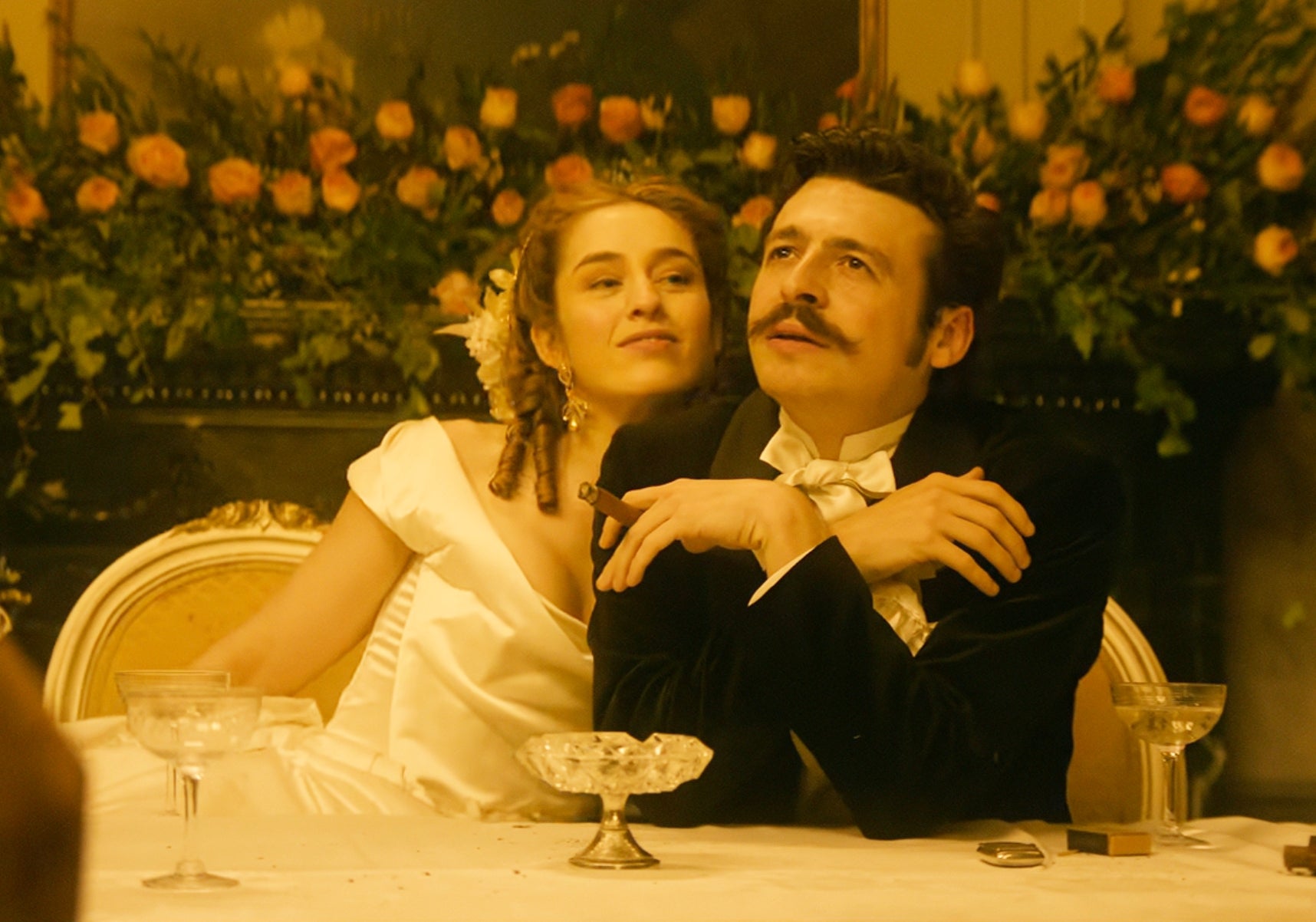 Two people in formal attire, with one in a white gown and the other in a black velvet suit with a bow tie, at an elegant dinner setting