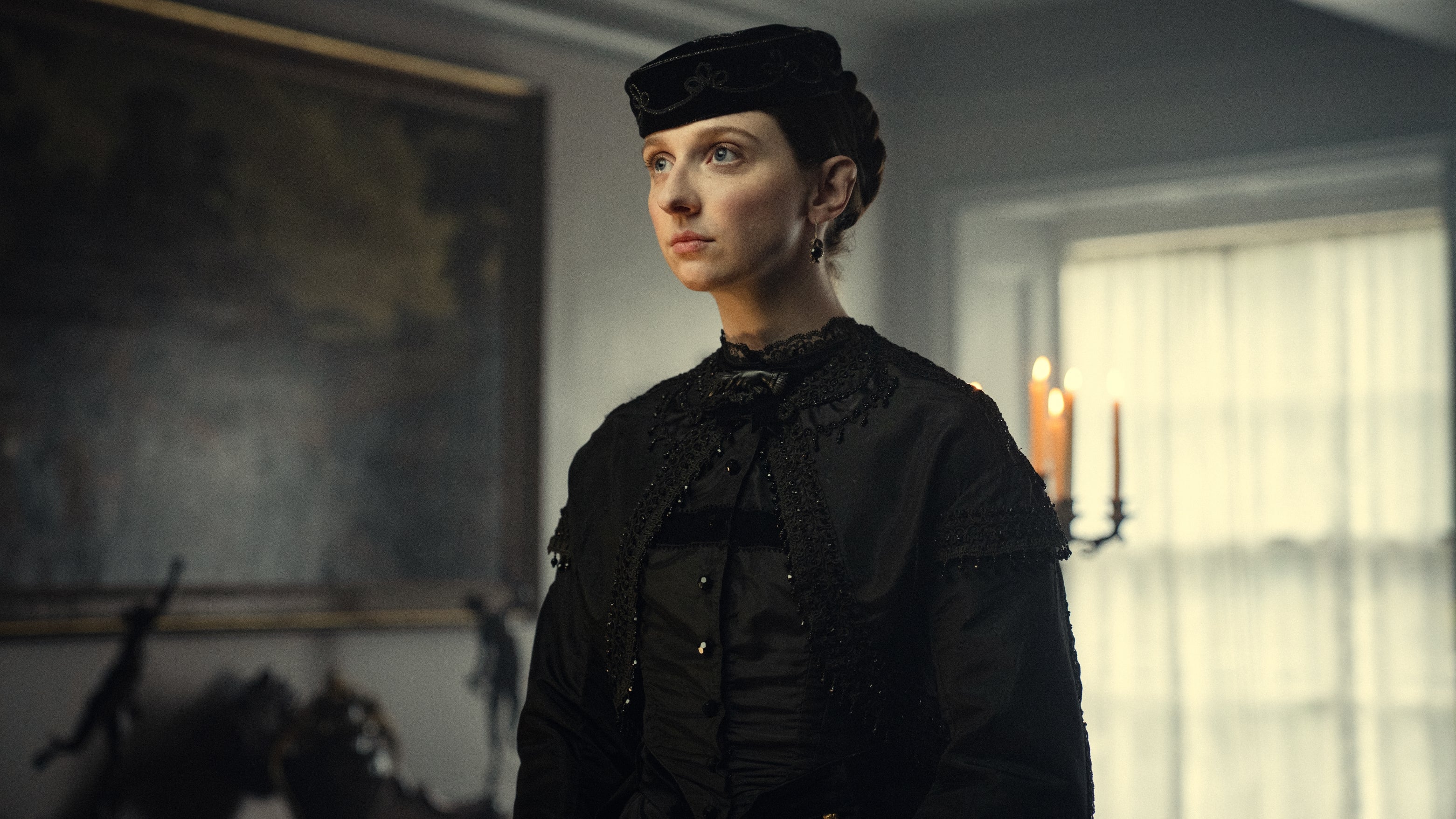 Person wearing Victorian-style black outfit with lace details, inside a dimly lit room, near a candelabrum and a painting
