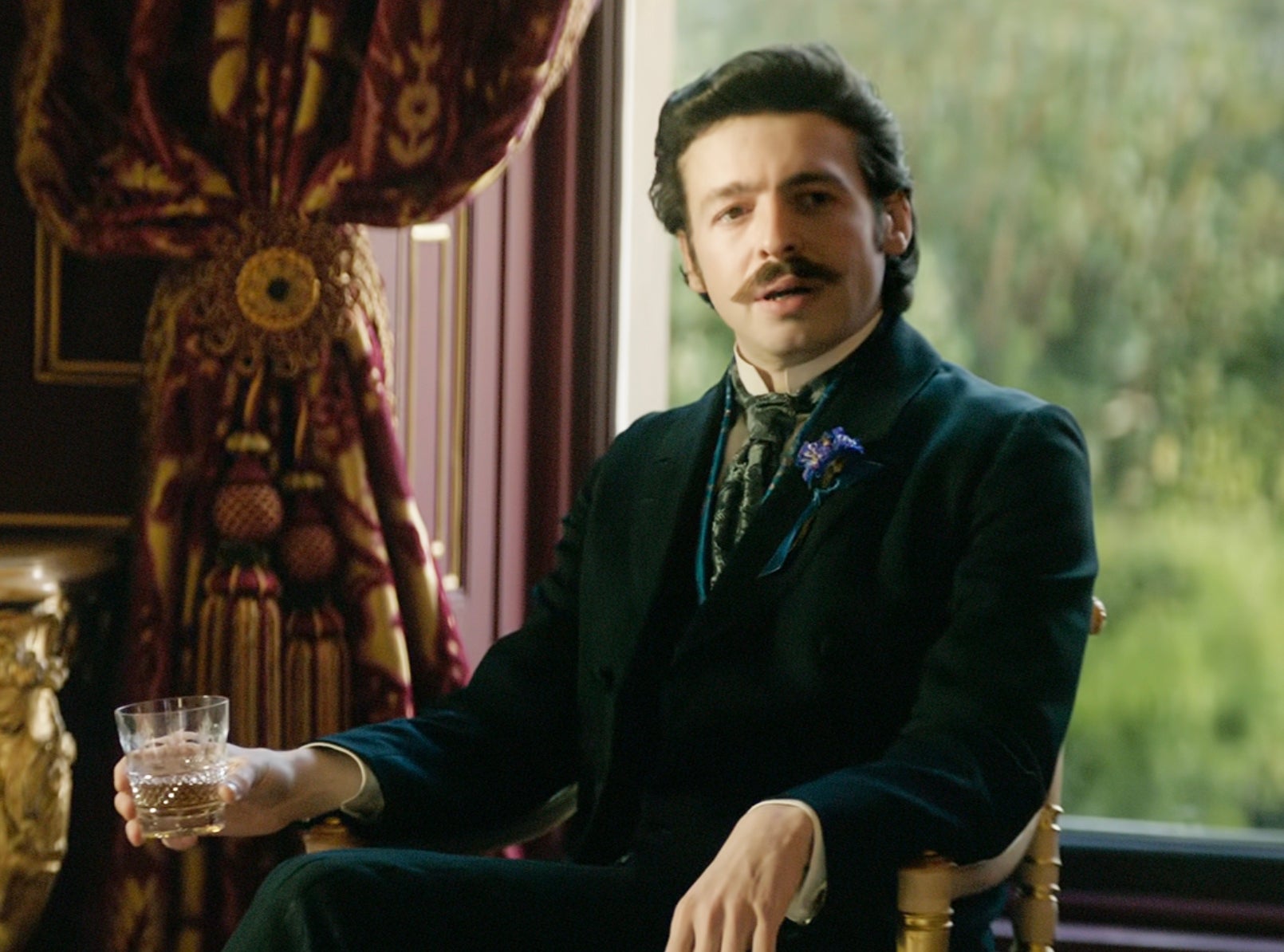 A person in period clothing holding a glass, seated in an elegant room, reflecting a historical or theatrical setting
