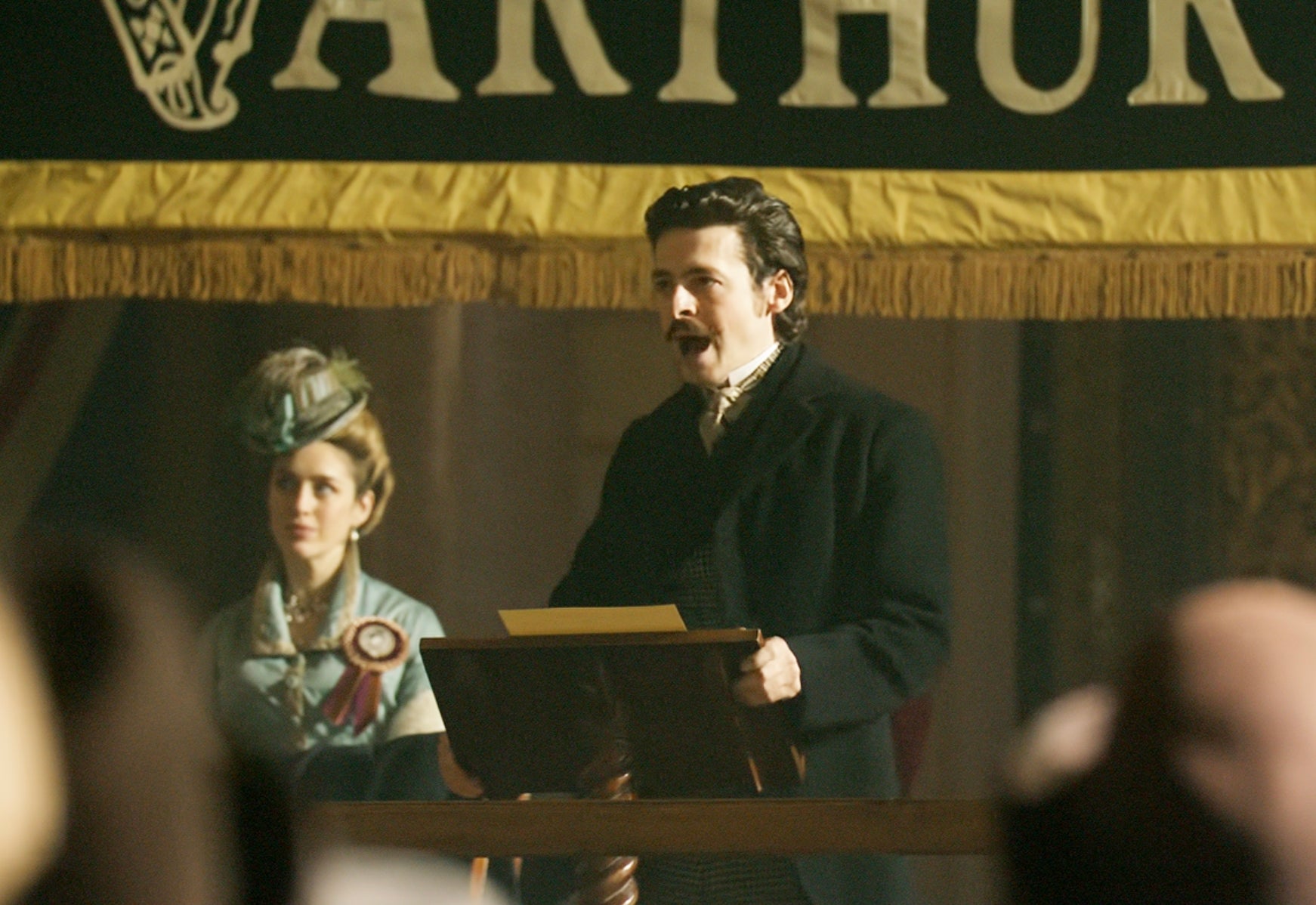 A man in period costume passionately speaks at a podium, with a woman in period attire standing behind him, in a historical setting
