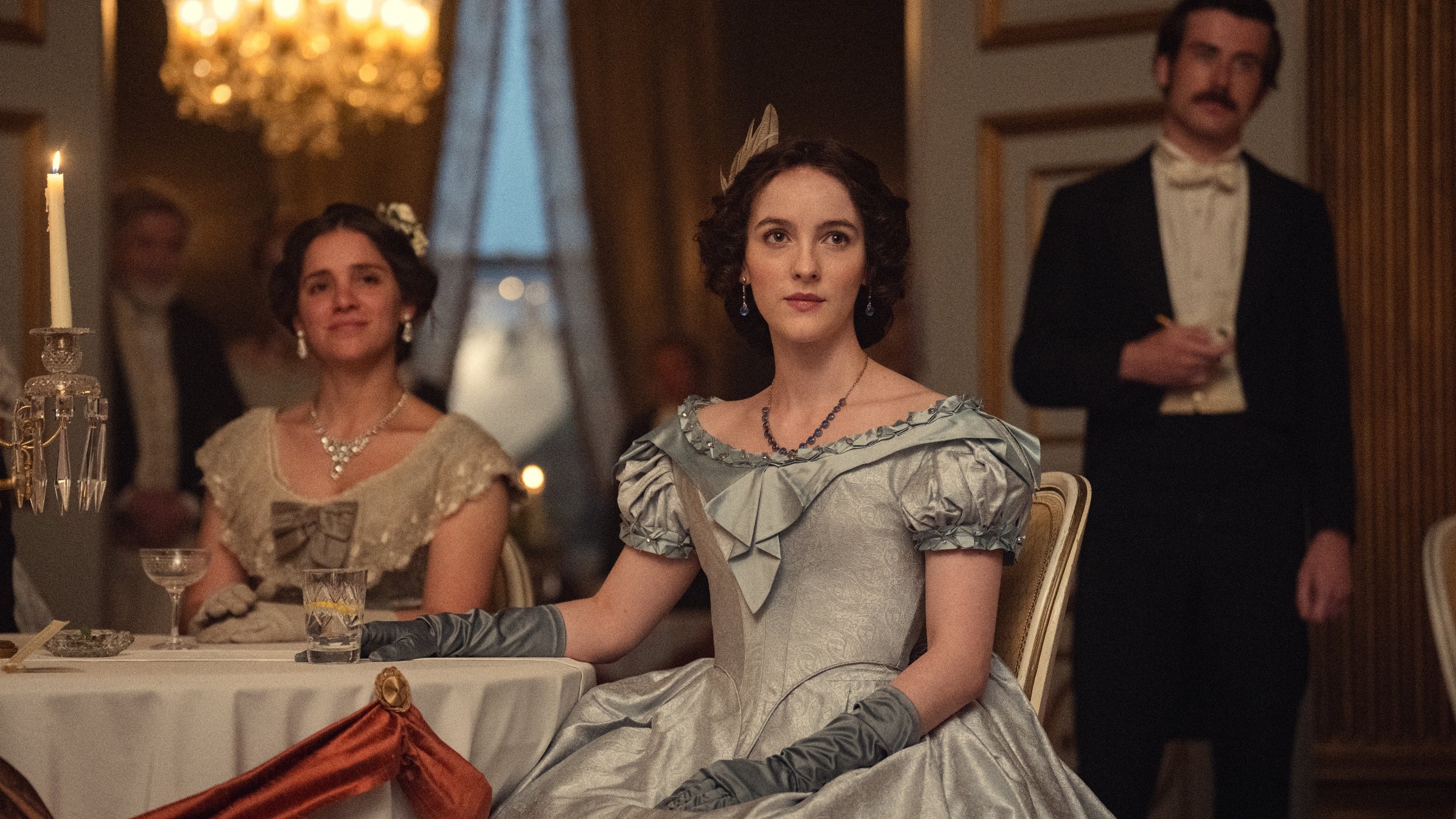 Actors in period-style dresses and formal attire sit elegantly at a table in a historical setting, conveying a vintage or period drama scene