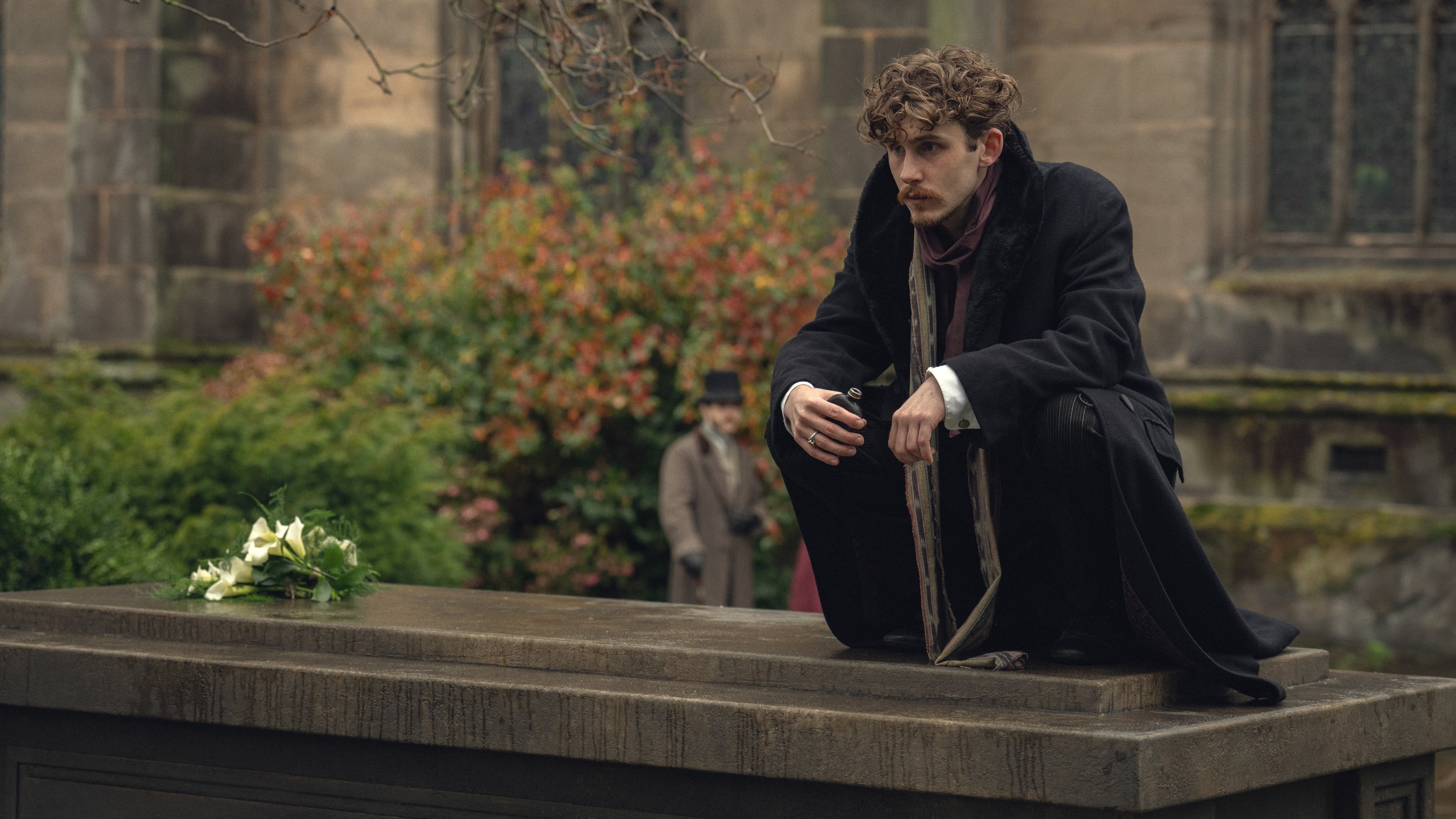 Man in period costume crouches on a gravestone, holding a pipe. A bouquet of white flowers lies nearby. Background shows a historical setting