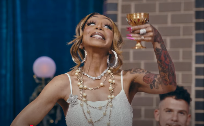 Person joyfully raising a gold goblet, wearing a sleeveless dress with layered necklaces and hoop earrings