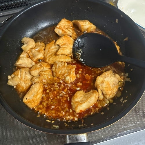 フライパンで調理中の鶏肉とソース。刻んだ野菜が混ざり、スプーンでかき混ぜられている料理の過程。