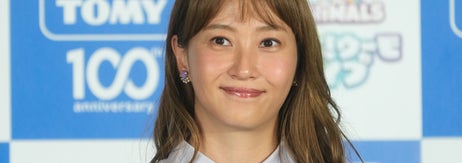 女性がイベントで、シンプルなシャツを着用し、カラフルなネックレスを身につけて微笑んでいます。バックにロゴがあります。