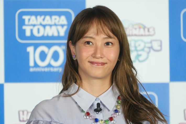 女性がイベントで、シンプルなシャツを着用し、カラフルなネックレスを身につけて微笑んでいます。バックにロゴがあります。